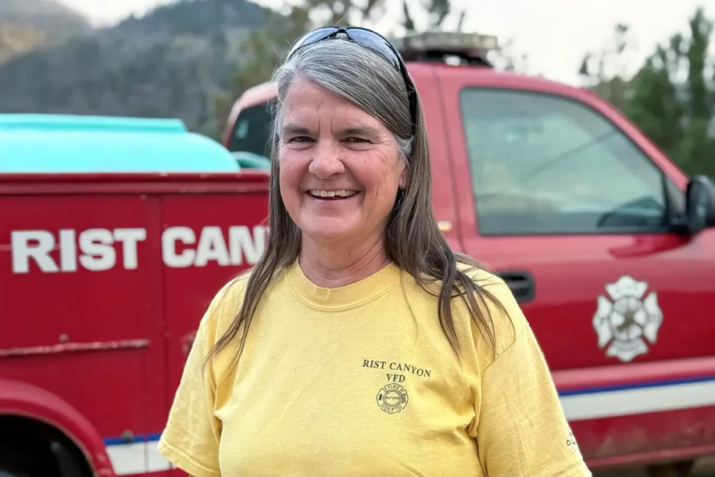 A woman in a yellow "Rist Canyon VFD" shirt stands smiling in front of a red fire department vehicle with trees and hills in the background.