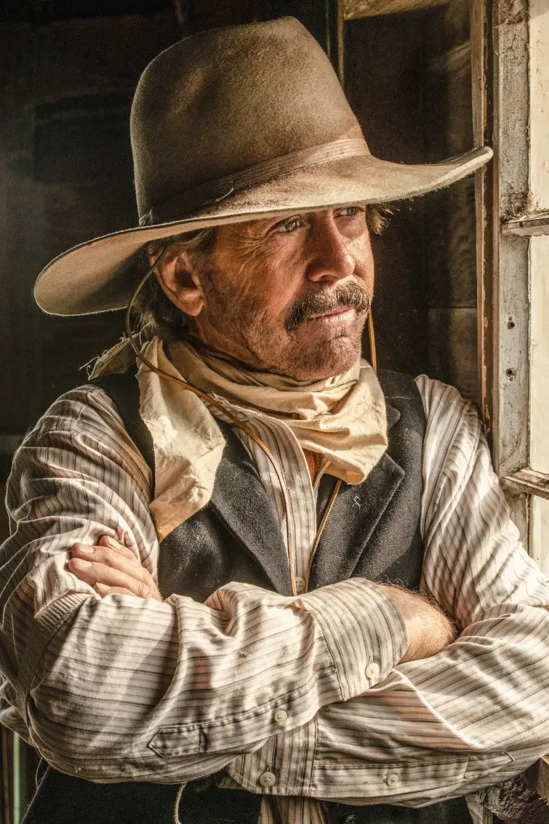 A man wearing a wide-brimmed cowboy hat, striped shirt, vest, and bandana stands with arms crossed, gazing thoughtfully out a window in warm, natural light.