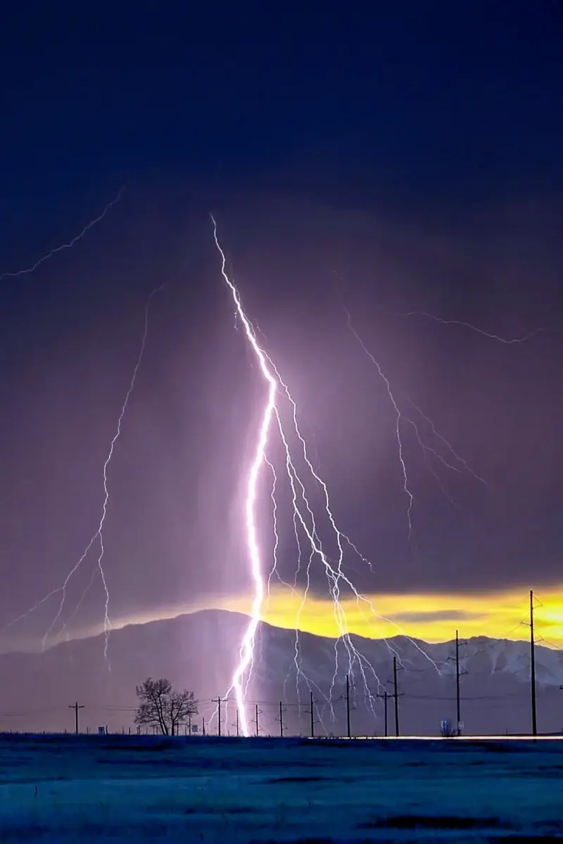 A powerful bolt of lightning strikes down from a dark, stormy sky over a mountain range at dusk, with a yellow glow on the horizon and silhouettes of utility poles and trees in the foreground.