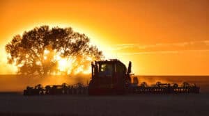 Silhouette of a tractor plowing a field at sunset, with a large tree and orange sky in the background. Dust is visible in the air.