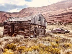 Old wooden cabin and abandoned car in a dry, grassy landscape with red rocky cliffs under a cloudy sky.