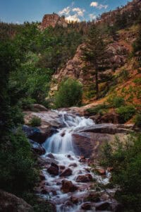 A cascading waterfall flows over rocks surrounded by lush greenery and trees, with a rocky hillside and blue sky in the background.