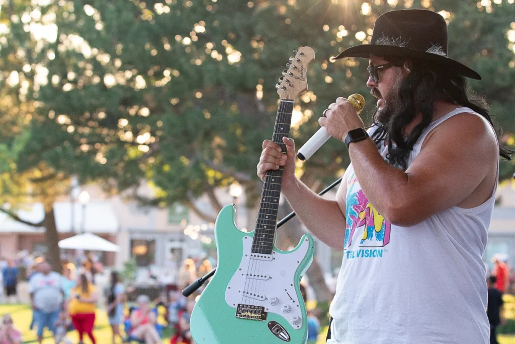 A man wearing a black hat and sunglasses sings into a microphone while holding a mint green electric guitar at an outdoor event.