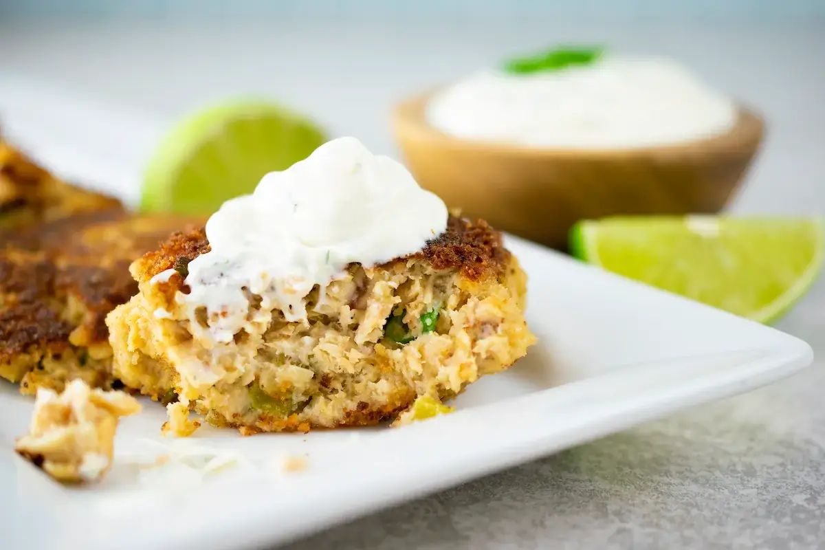 A sliced fish cake topped with sour cream on a white plate, accompanied by lime wedges and a small bowl of creamy sauce in the background.