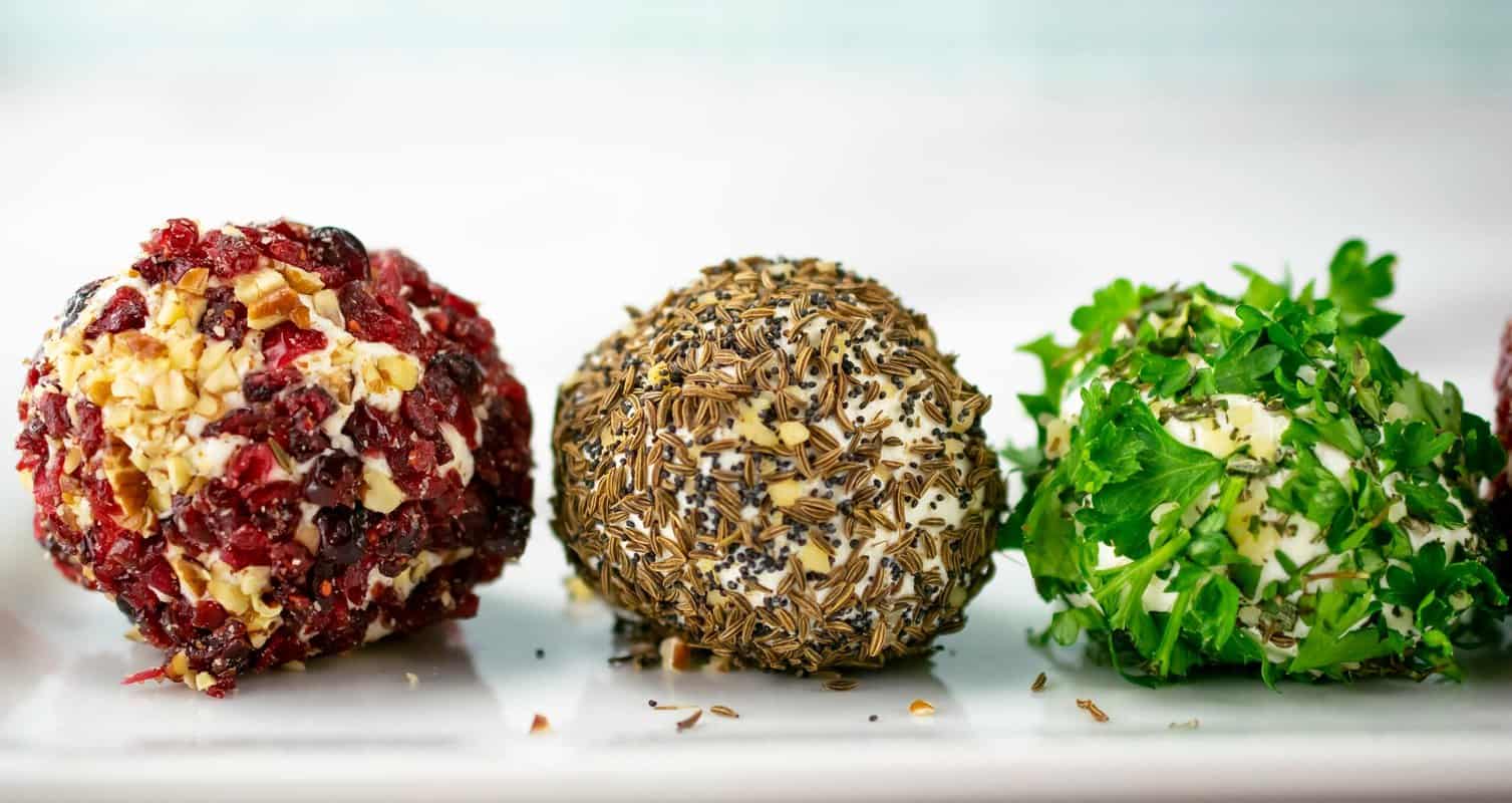 Three cheese balls coated with chopped cranberries and nuts, seeds, and parsley, arranged on a white plate.