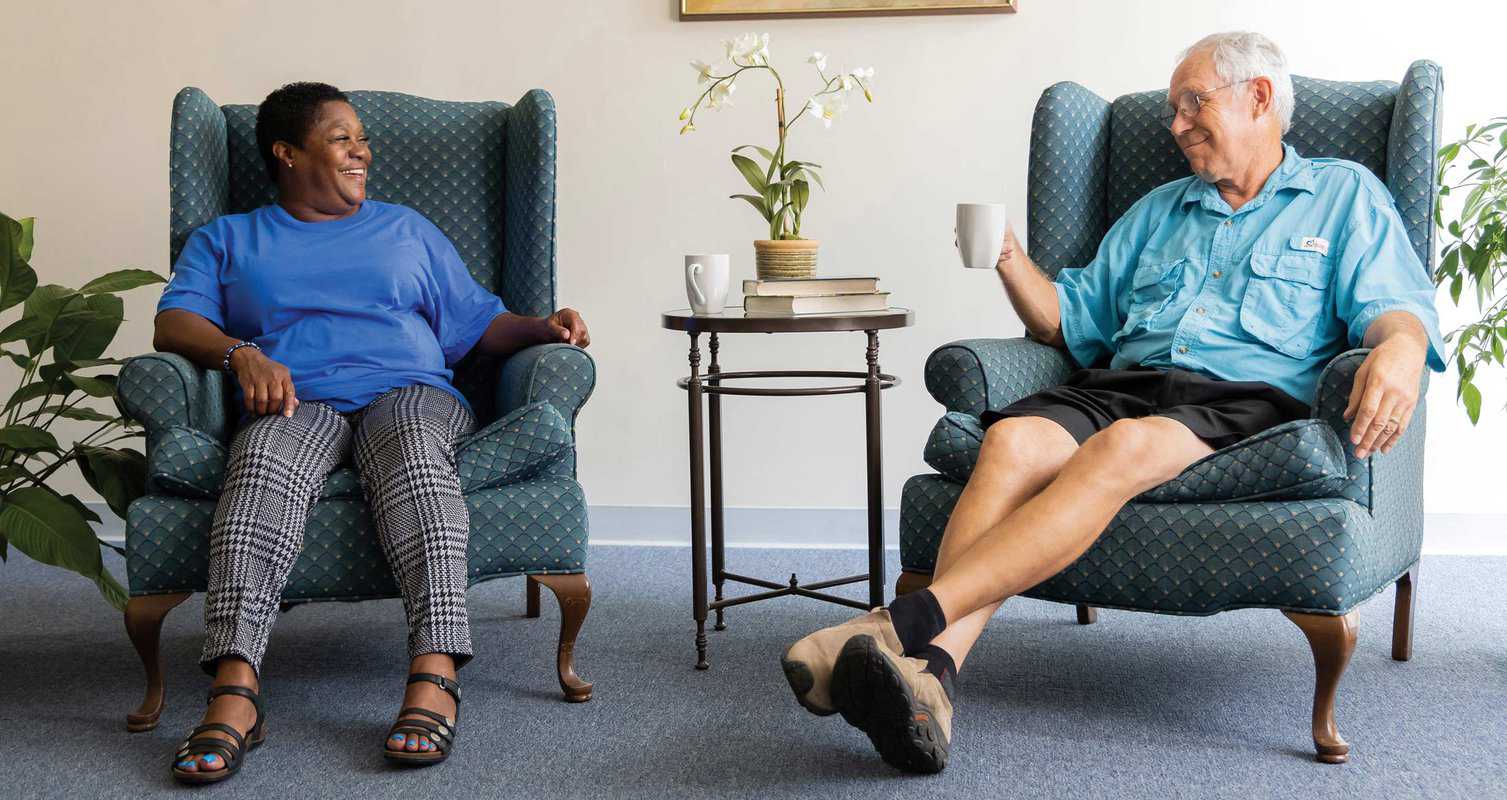 Two people sitting in armchairs, smiling at each other. A small table with books, a plant, and coffee cups is between them.