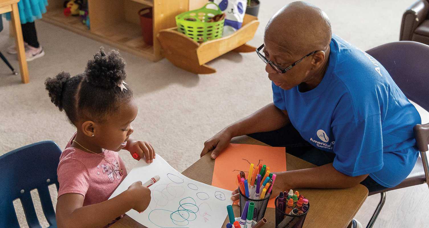 A young girl draws with crayons while an adult sits beside her, observing. Various art supplies are on the table.