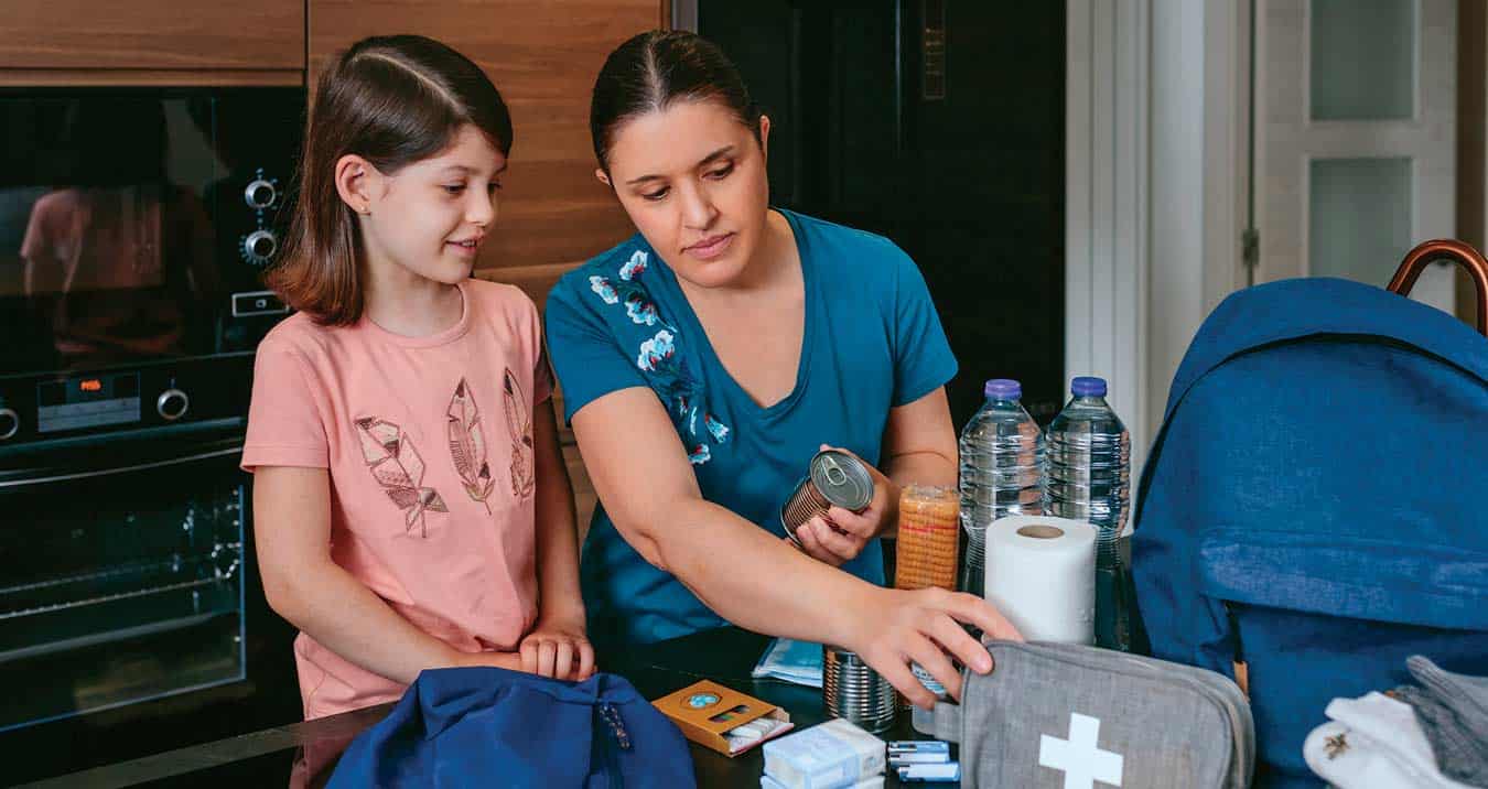 Woman and child preparing an emergency kit on a kitchen table with canned food, bottled water, a first aid kit, and other supplies.