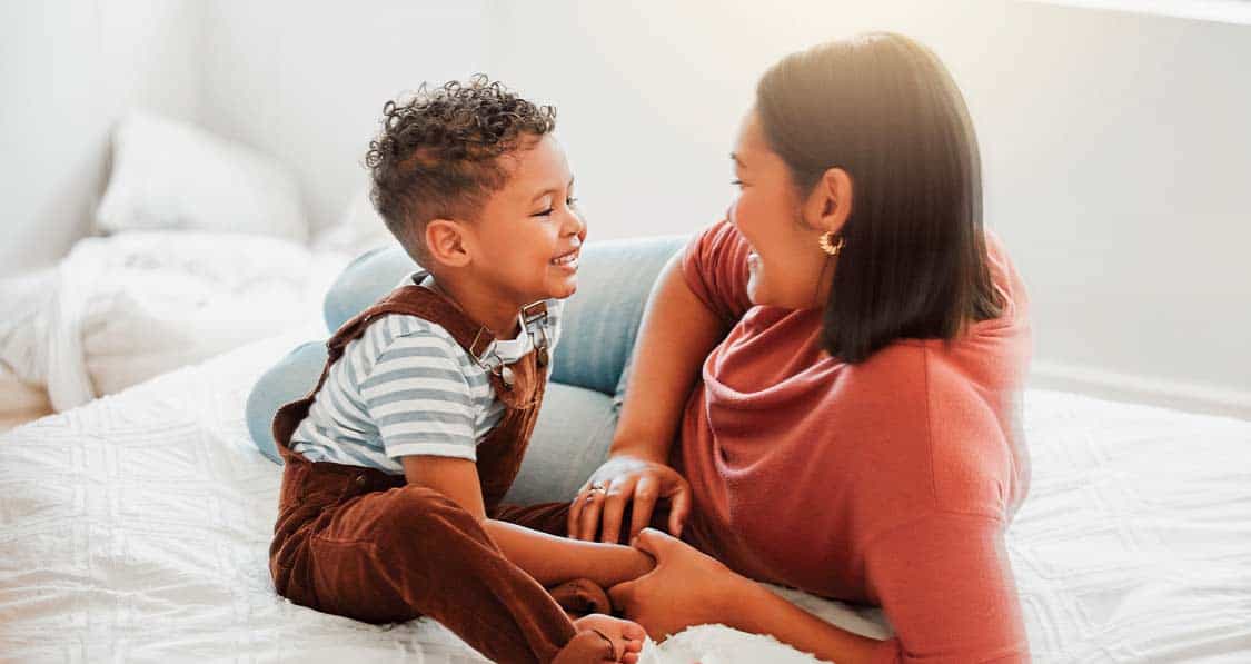 Woman and child sitting on a bed, smiling at each other in a brightly lit room.