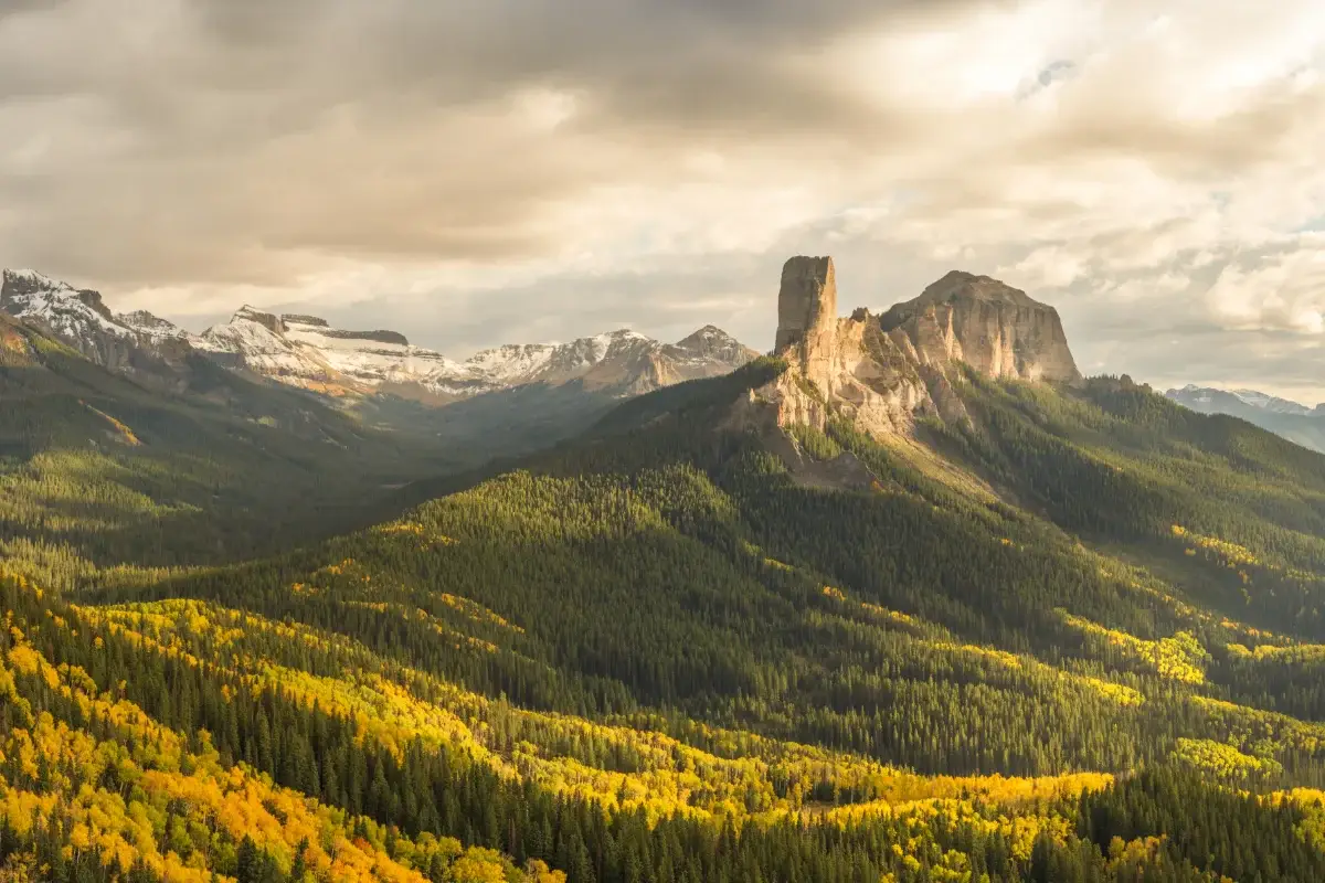 Sunlight filters through clouds, illuminating a dramatic rocky peak surrounded by dense forests and rolling hills with trees in shades of green and yellow, set against a backdrop of distant snow-capped mountains.