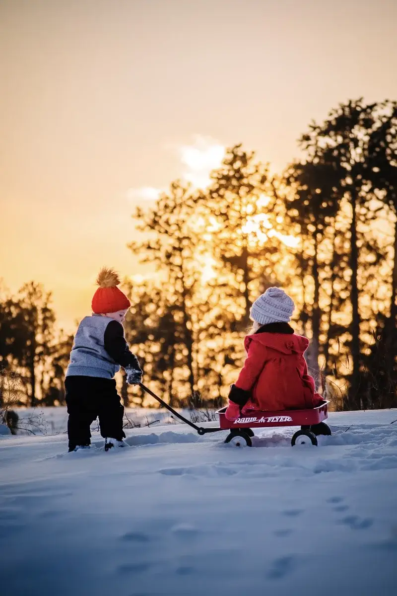 Two young children in winter clothing play outdoors at sunset; one pulls a red wagon with the other child inside across snow, with tall trees and a golden sky in the background.