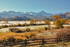 Scenic autumn landscape with snow-dusted fields, yellow and orange trees, and mountain range under a clear sky. Foreground features a wooden fence.