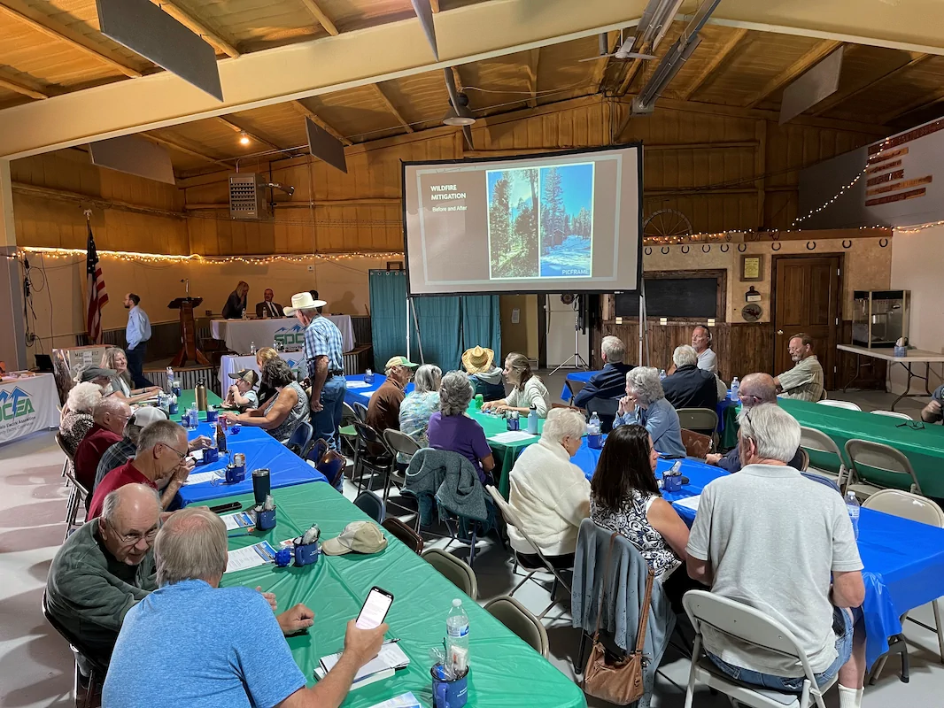 People seated at tables in a community hall watch a presentation projected on a screen at the front, with speakers standing near a podium.