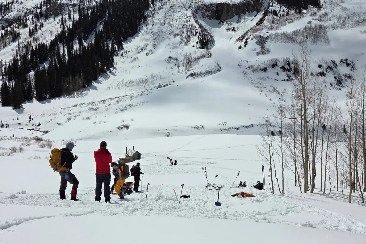 The Next Generation of Search and Rescue A group of people with backpacks and equipment stand on a snowy slope, with trees and mountains covered in snow in the background.