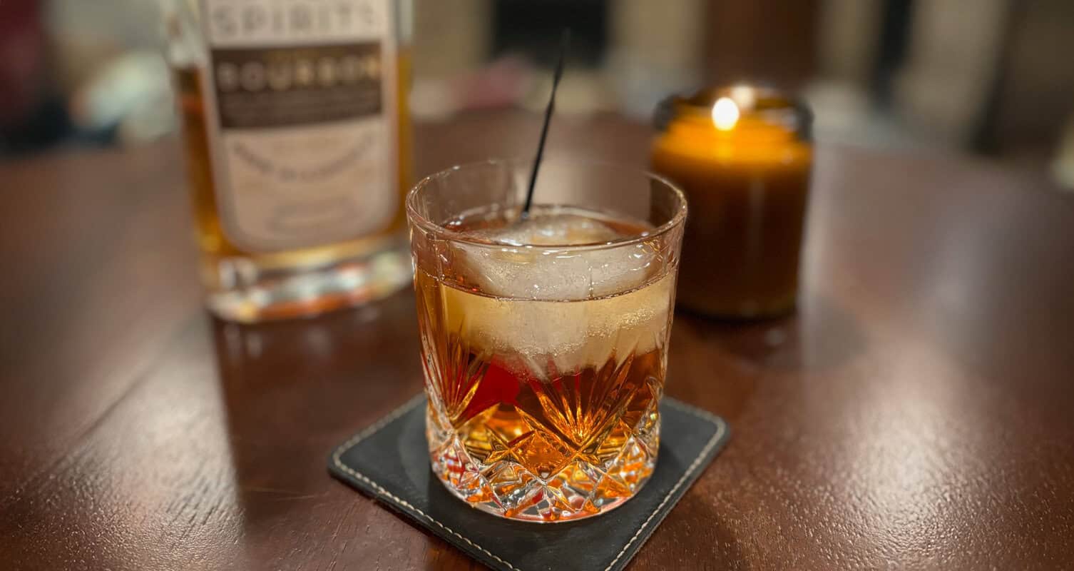 A glass of whiskey with ice on a coaster, next to an unlit candle and a bottle of bourbon, on a wooden table.