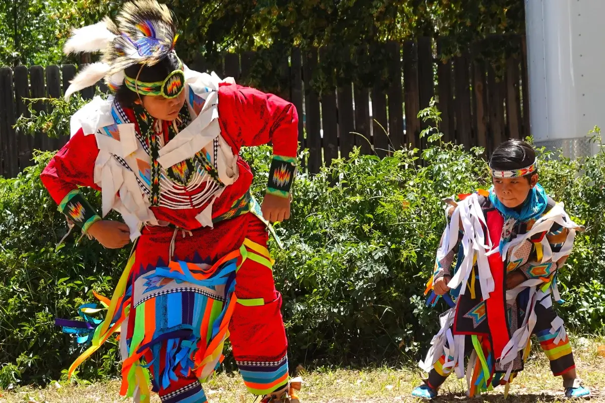 Two people in colorful, traditional Indigenous regalia dance outdoors on grass, surrounded by greenery and sunlight. The larger dancer wears red, while the smaller dancer wears darker patterns. Both have ribbons and beadwork on their outfits.