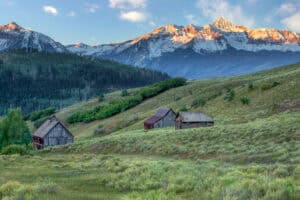 Mountain landscape with three rustic wooden cabins on grassy hills in the foreground and snow-capped peaks in the background under a cloudy sky.