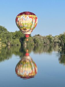 A colorful hot air balloon floats above a calm lake, reflecting perfectly in the water. Trees line the background under a clear blue sky.