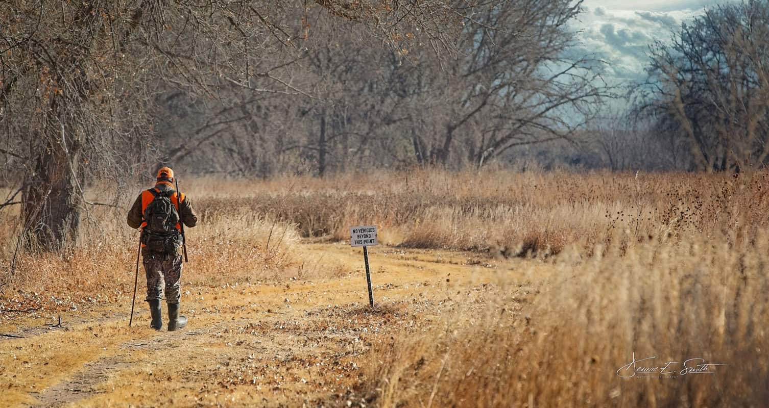 Hunter in camouflage and orange vest walks down a dirt path in a dry, wooded area under a cloudy sky. Sign nearby reads "Vehicles Prohibited Beyond This Point.