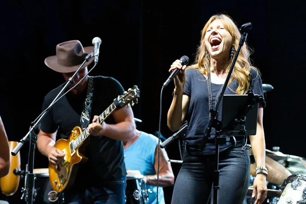 A woman passionately sings into a microphone while a man plays electric guitar on stage during a live music performance.