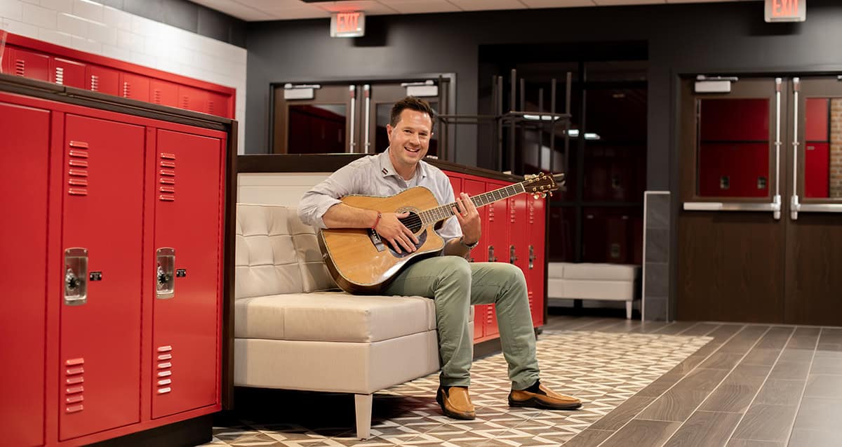 A person sitting on a cushioned bench plays an acoustic guitar in a room with red lockers and tiled floor.