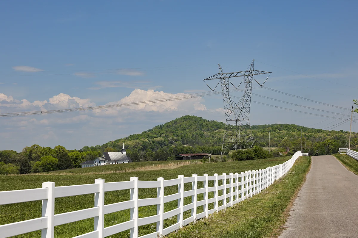 A country road lined with a white fence runs beside a grassy field. Power lines and a tall transmission tower stand near a green, tree-covered hill under a blue sky with scattered clouds. A white barn is in the distance.