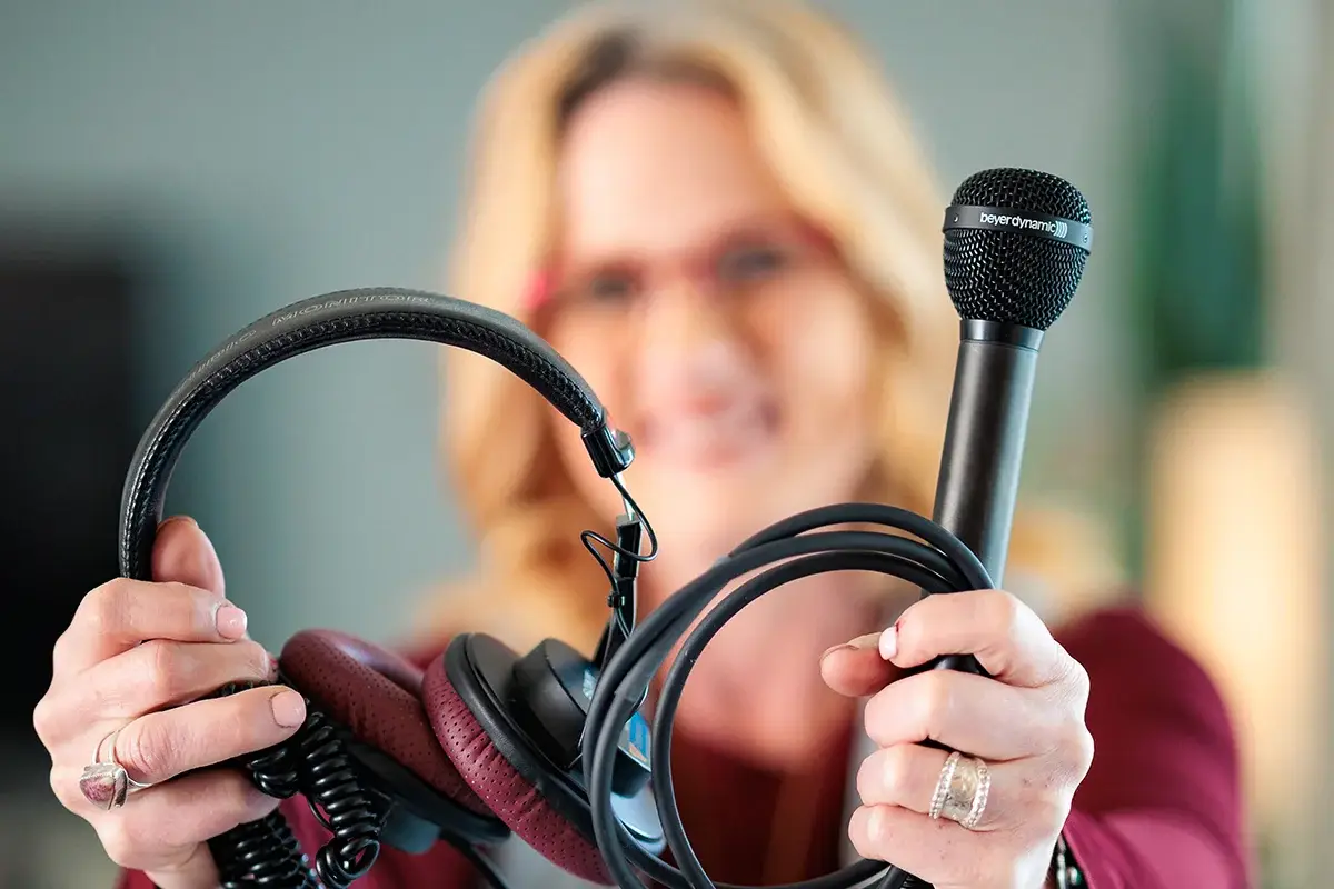 A woman with blonde hair holds a pair of headphones in one hand and a black microphone in the other, both extended toward the camera. Her face is out of focus in the background.