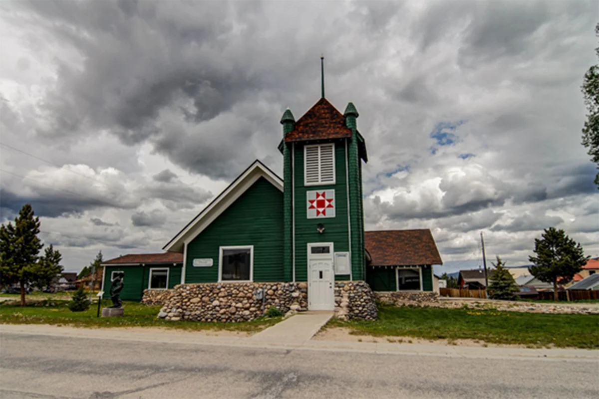 Giving Space A small green church with a stone foundation and a tall central steeple stands under a cloudy sky. A red and white star pattern decorates the steeple. Trees and grass surround the building.