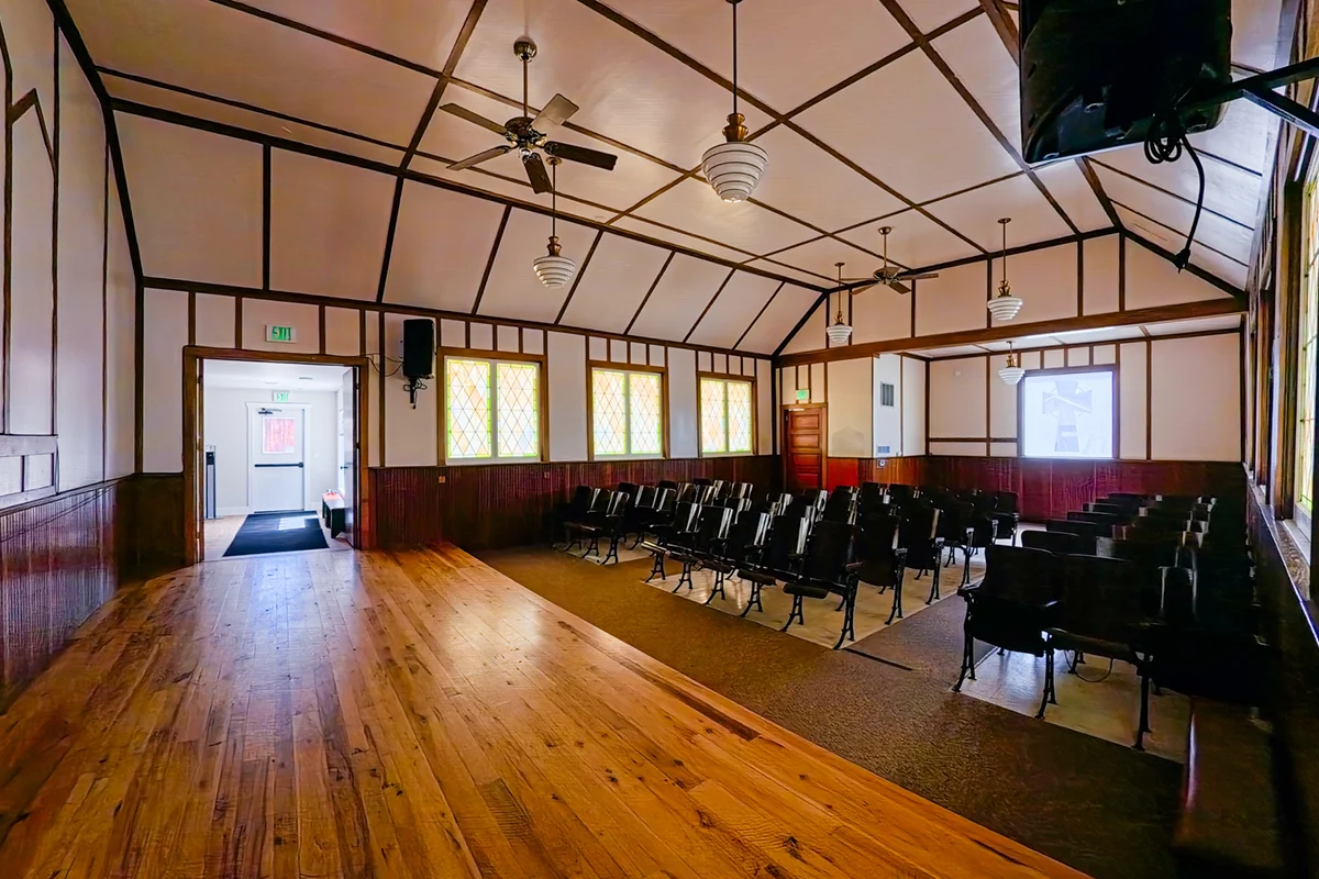 Giving Space Spacious room with wooden floors, rows of black chairs, high ceiling with fans and lights, large windows with yellow stained glass, and an open doorway leading to a hallway.
