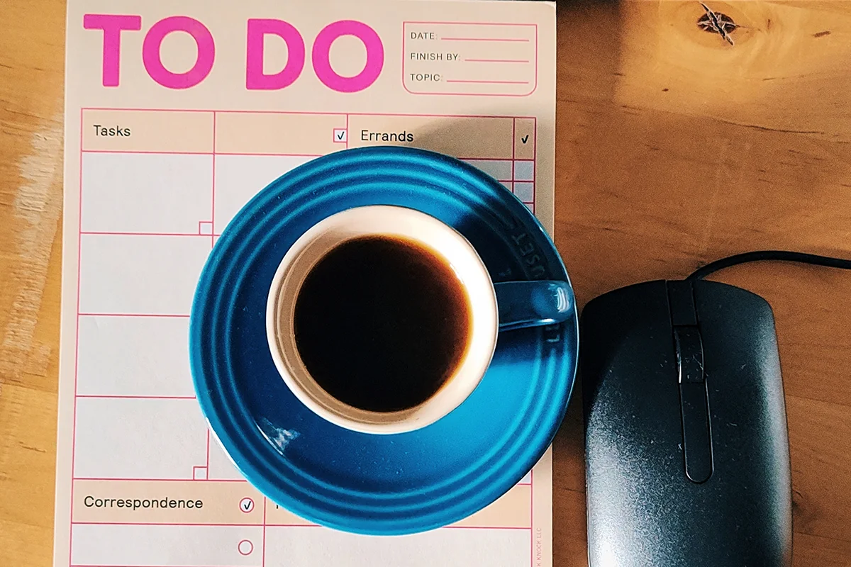 Rural Sleep Reset A blue coffee cup on a to-do list pad with columns for tasks, errands, and correspondence, next to a black computer mouse on a wooden desk.