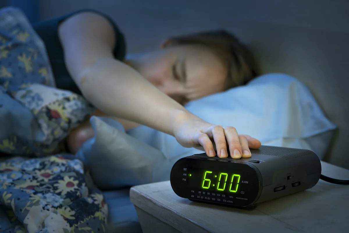 A person lying in bed reaches out to turn off a digital alarm clock showing 6:00 AM. The room is dimly lit and the person appears to be waking up, partially covered with a floral-patterned blanket.