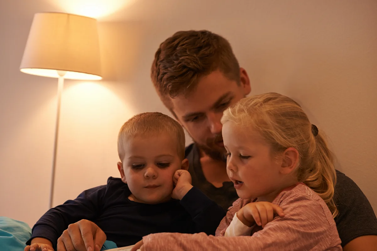 Rural Sleep Reset A man sits with two young children, reading together in a softly lit room with a lamp in the background. The children are engaged and listening closely.