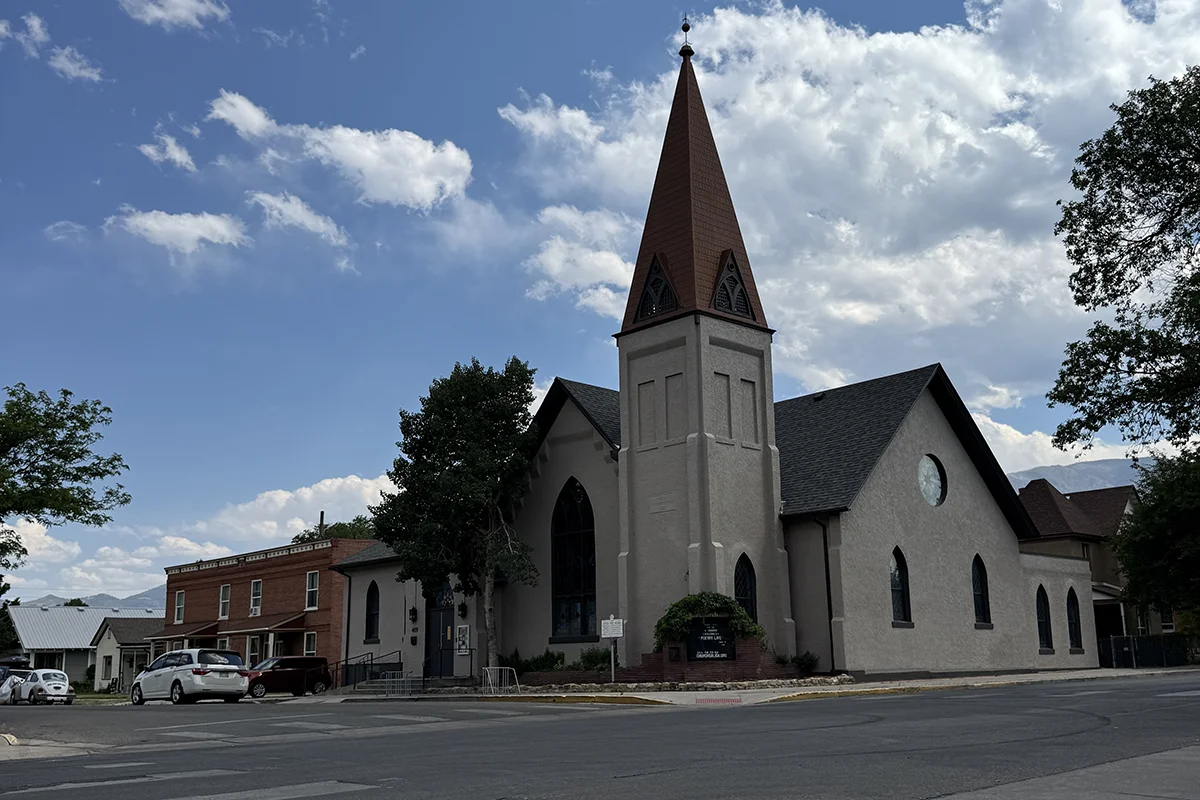 Giving Space A church with a tall steeple and large arched window stands on a street corner, surrounded by trees, cars, and nearby buildings under a partly cloudy sky.