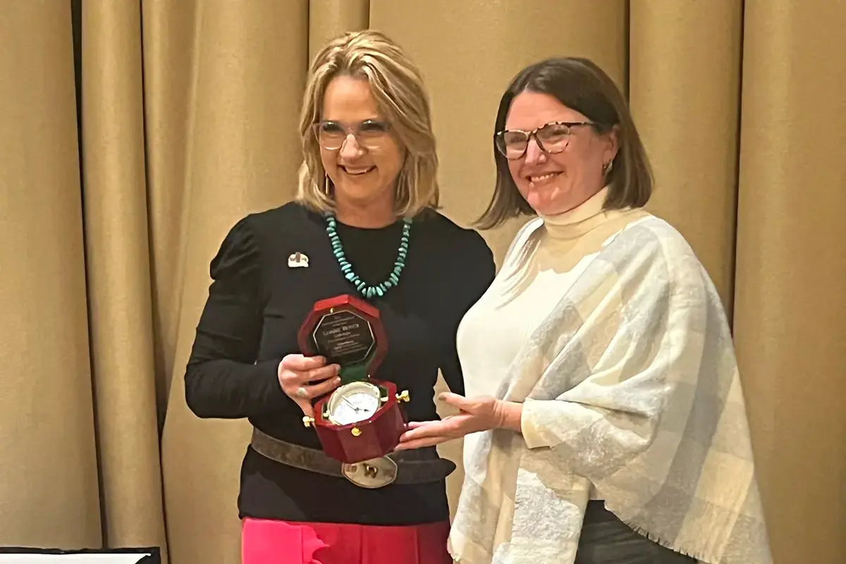 Two women stand in front of beige curtains, smiling. One woman holds an open wooden box displaying a clock and plaque, as the other woman gestures toward it. Both are wearing glasses and casual business attire.
