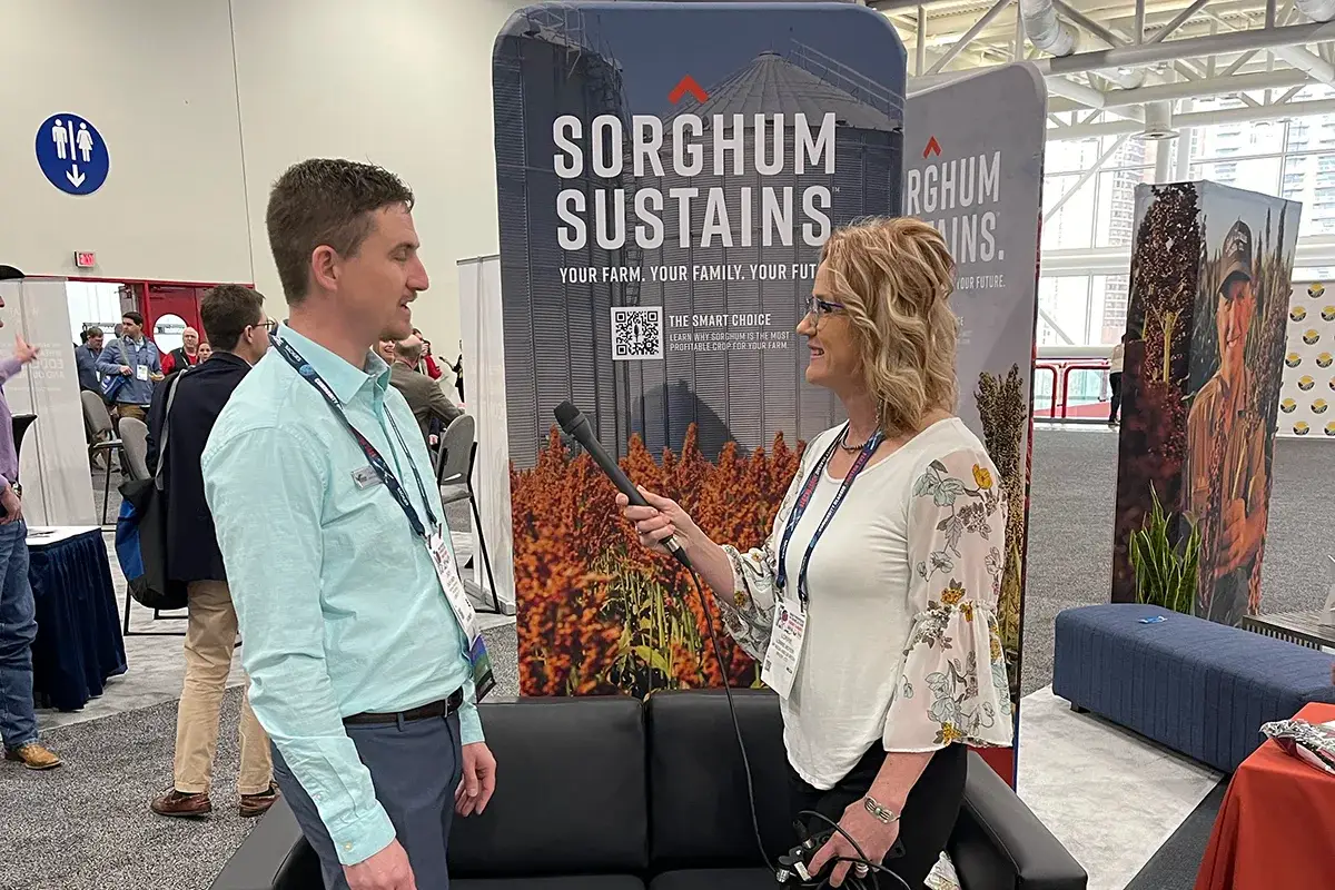 A woman interviews a man with a microphone at an event booth featuring a display that reads "SORGHUM SUSTAINS" with images of sorghum plants and a silo in the background. Other attendees and booths are visible behind them.
