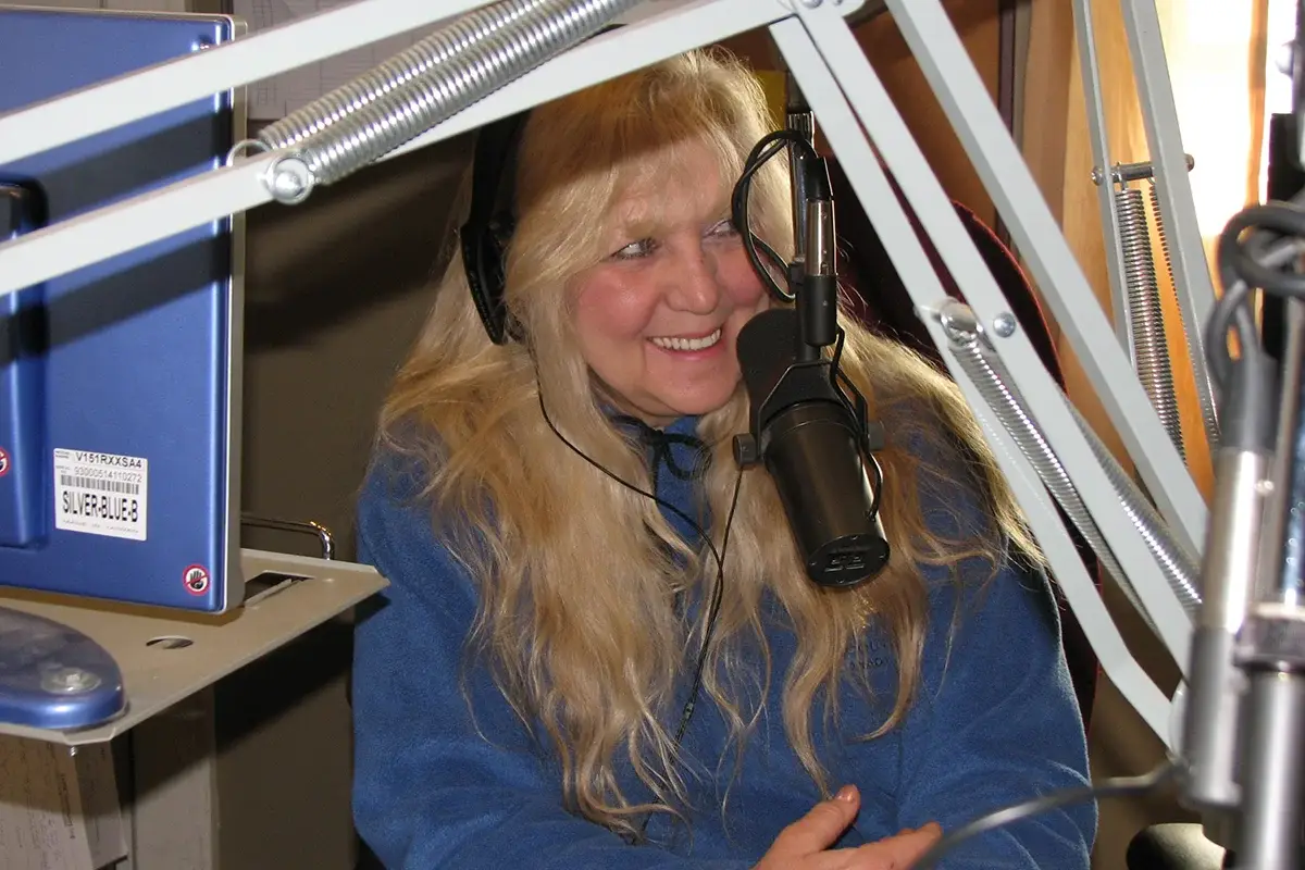 A woman with long blonde hair sits at a radio station, smiling as she speaks into a large microphone. She is wearing headphones and a blue fleece jacket, surrounded by studio equipment.
