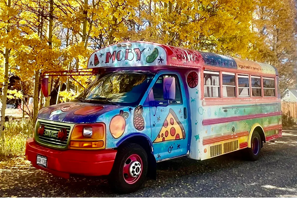 Fulfilling a Community's Heart A colorful, decorated bus with pizza and fruit art is parked on a road, surrounded by vibrant yellow autumn trees.
