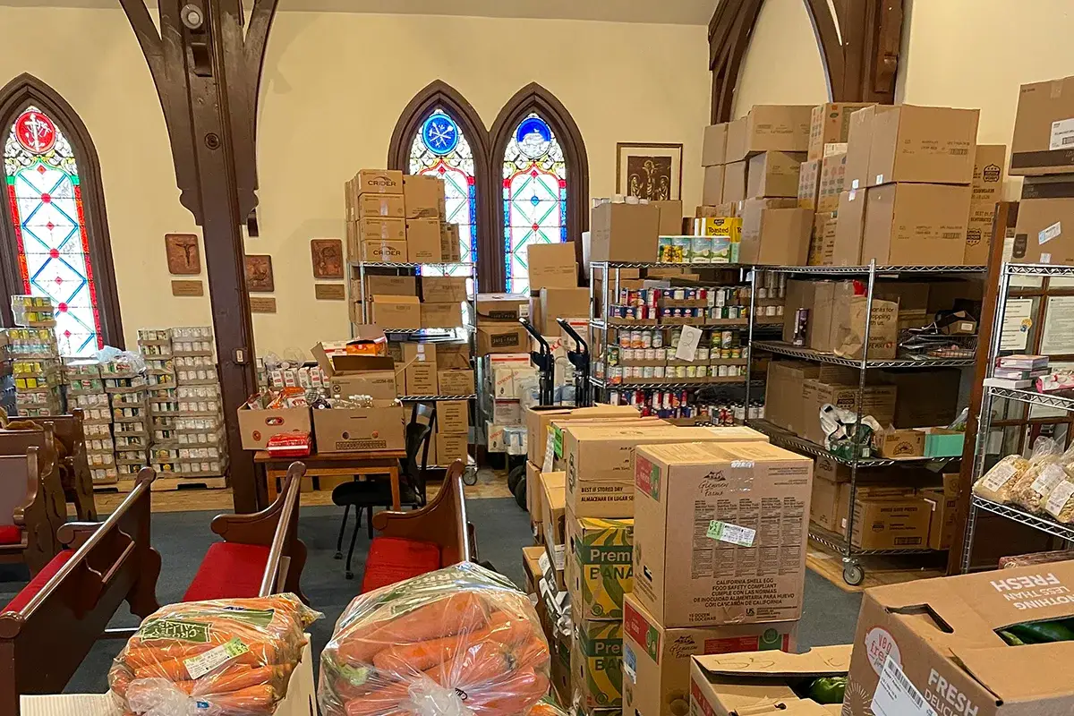 Fulfilling a Community's Heart A church room filled with shelves and stacks of boxes, bags of carrots, canned goods, and other food items, indicating a food pantry or donation center. Stained glass windows are visible in the background.