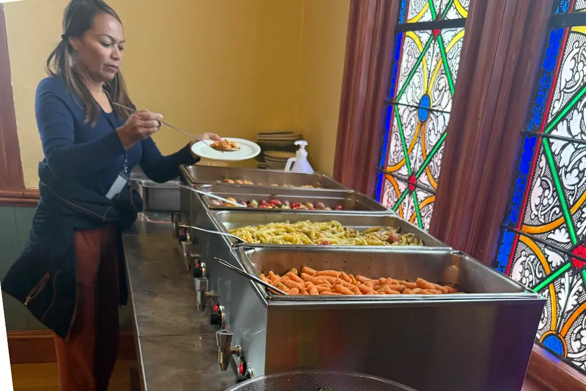 Fulfilling a Community's Heart A woman serves herself food from a buffet with trays of pasta, carrots, and other dishes in front of stained glass windows.