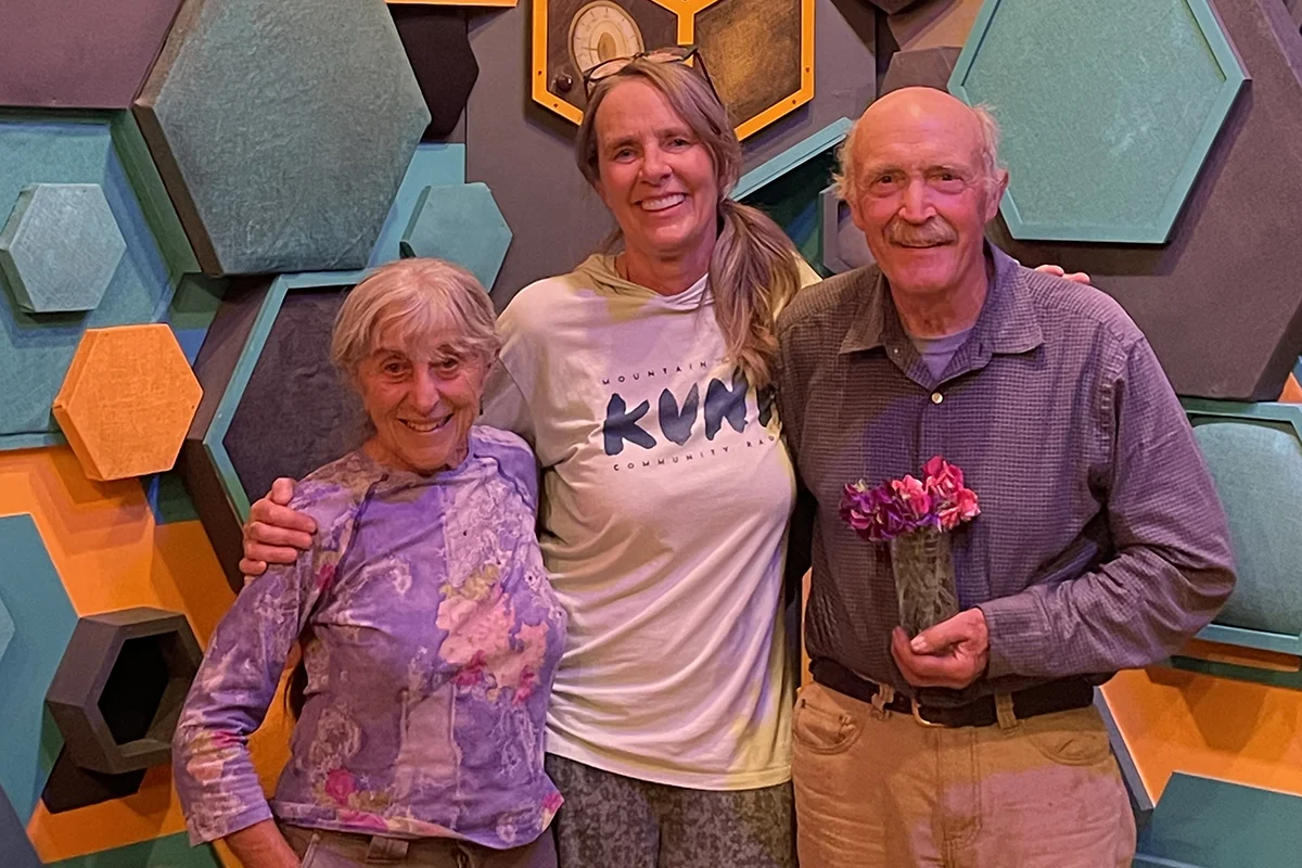 Three adults smile together in front of a colorful geometric wall; the person in the middle has their arms around the two older adults, one of whom holds a small vase of flowers.