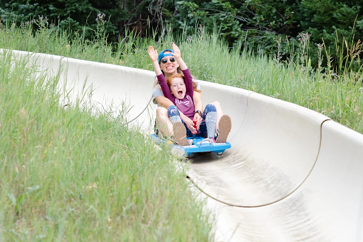 An adult and a child, both smiling with their arms raised, ride together on a blue sled down a curved outdoor slide surrounded by green grass and trees.