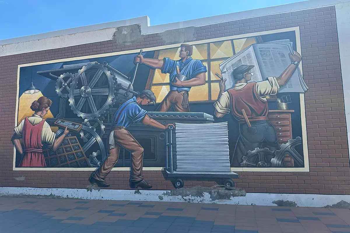 A mural on a brick wall depicts four workers operating a large printing press, with one holding a large sheet of paper and others engaged in various tasks, set against a background of machinery and tall windows.