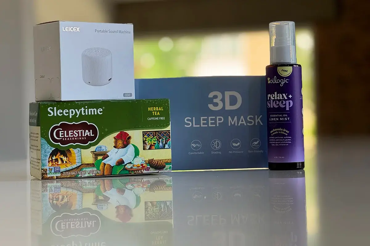 A box of Celestial Seasonings Sleepytime Herbal Tea, a LEICEX portable sound machine, a 3D sleep mask, and a bottle of Biologic relax+ sleep lavender mist arranged on a reflective surface.