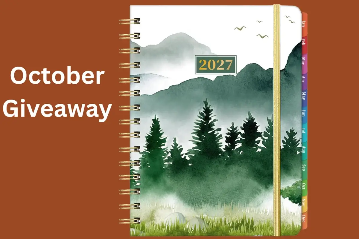 Spiral-bound 2027 planner with a forest and mountain cover, colorful side tabs, and gold elastic band. Text reads “October Giveaway” on a brown background.