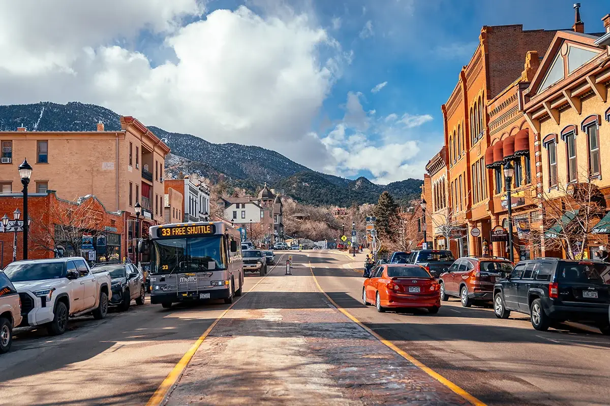 A small town street with parked cars, a bus labeled "33 FREE SHUTTLE," brick buildings on both sides, and snow-capped mountains in the background under a partly cloudy sky.