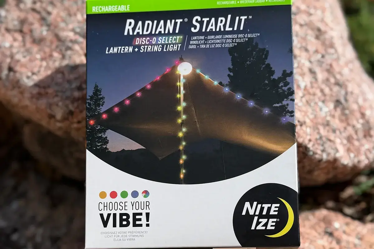 A box of Nite Ize Radiant StarLit lantern and string lights is shown outdoors on rocks, featuring multicolored lights strung over a tent with "Choose Your Vibe!" and product details on the packaging.