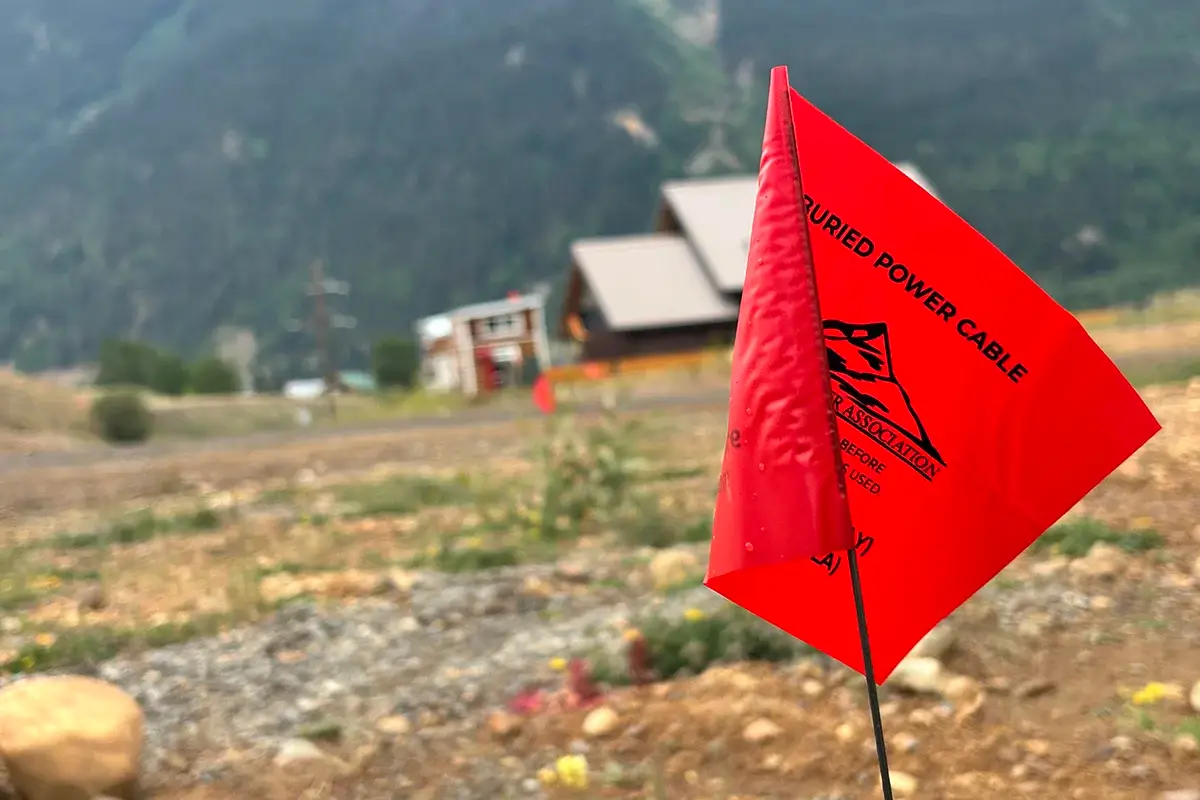 A close-up of a red warning flag labeled "BURIED POWER CABLE" in a rocky, rural area with a house and mountains blurred in the background.