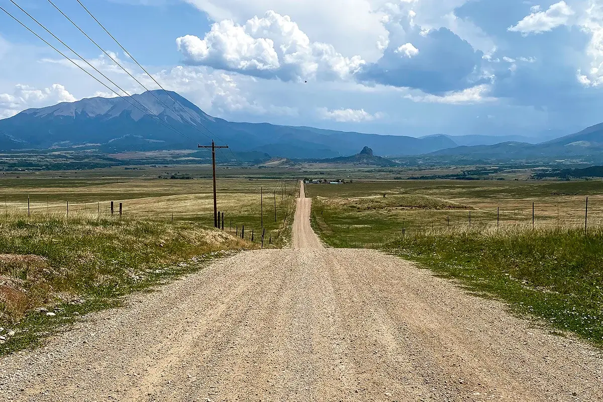 A long gravel road stretches straight through open fields toward distant mountains under a partly cloudy sky, with power lines running along one side.