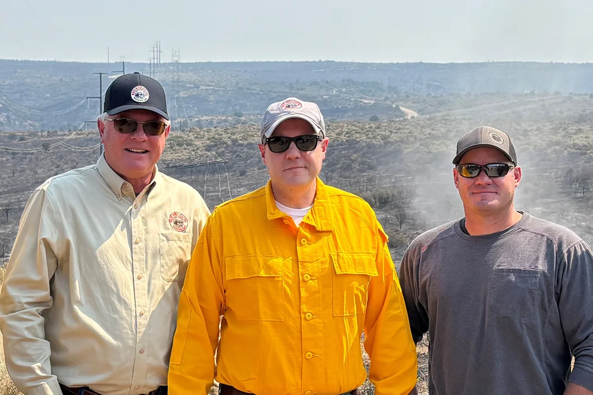 Three men wearing sunglasses and hats stand outdoors in front of a smoky, hilly landscape. One wears a yellow shirt, the other two wear light-colored and gray shirts. Power lines and brush are visible in the background.