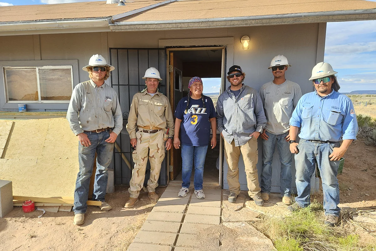 Six people stand smiling outside a house under construction. Five wear hard hats and work clothes, one person in the center wears a blue shirt and bandana. The scene appears sunny and they stand on a dirt path.